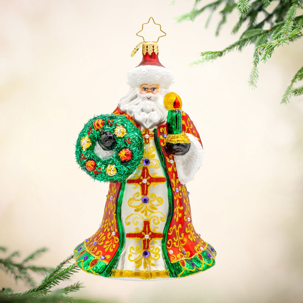 This stunning ornament showcases Santa in a resplendent red and gold robe, hand-painted with intricate designs and finished with shimmering trim. Carrying a glowing green candle and a wreath adorned with shiny holly and colorful ornaments. View of front.