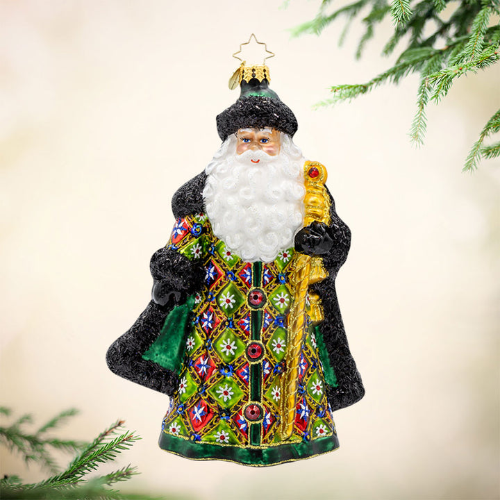 The Opulent Noel ornament. Santa is dressed in a vibrant green satin coat covered in a playful patchwork of colors and glittery designs. With his shiny gold staff, sparkling red gems, and fluffy tinsel fur trim. View of front. 