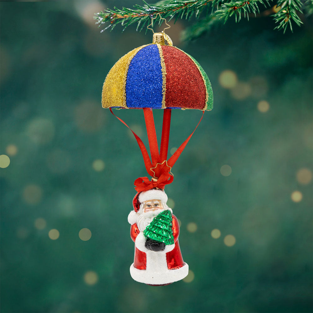 Tidings from Above Santa ornament. This ornament features Santa soaring through the air with a colorful parachute, his tiny Christmas tree in hand. View of front.